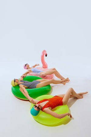 Three women lying on brightly colored inflatable pool floats, dressed in stylish swimsuits and swim...