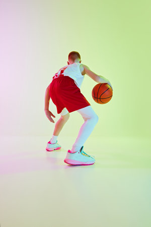 Dribbling. Back view image of boy in uniform, playing basketball game, training against gradient background in neon lightの写真素材