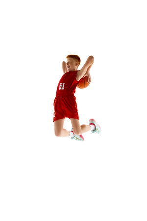 Full-length dynamic image of teen boy in red uniform, playing basketball, throwing all in jump isolated on white studio backgroundの写真素材