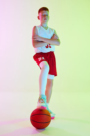 Motivated player. Full-length image of teen boy in uniform posing with basketball ball against gradient background in neon lightの写真素材