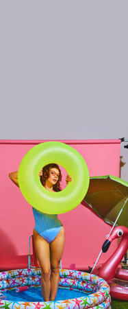 Joyful young woman in striped, retro swimsuit stands in inflatable pool, playfully holding...