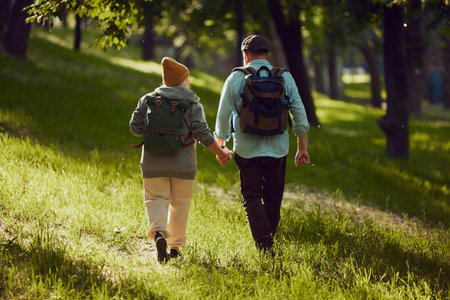Back view of elderly couple holding hands and enjoying leisurely hike in sunny green forest, wearing comfortable clothes and backpacksの写真素材