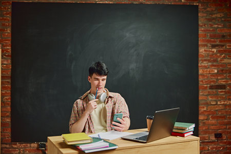 Young guy, student sitting in classroom at desk with laptop, books, coffee and chalkboard behind....