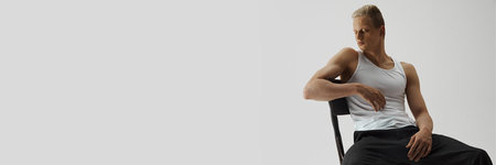 Young handsome man with muscular body posing on chair isolated on grey studio background. Confidence...