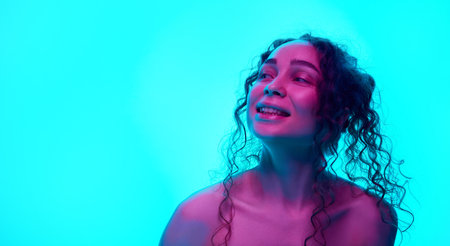 Happy, smiling, young woman with curly hair posing against cyan background in neon light. Spotless, well-kept skinの写真素材