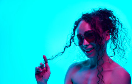 Portrait of smiling young woman with bare shoulder, curly hair in ponytail, posing in sunglasses against cyan background in neonの写真素材