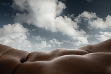 Beauty of cloud formations and human body curves. Close-up of tanned male muscular body forming natural landscape with cloudy sky on background.の写真素材