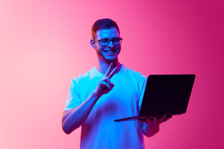 Smiling young man in glasses and white t-shirt holding laptop and having online video call with friends against pink background in neon lightの写真素材