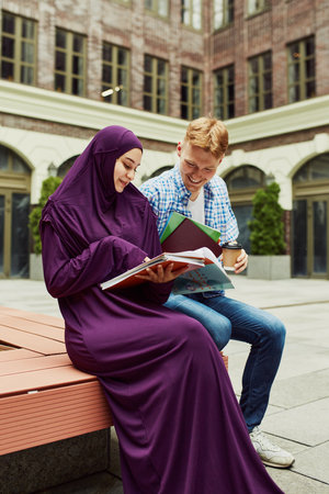 Smiling friends, students, Muslim girl and Caucasian boy sitting outside the campus and preparing for lesson together.の写真素材
