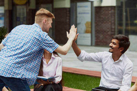 Hello. Caucasian young guy giving high five to Indian friend. Multicultural students meeting outdoors the campus for common studyingの写真素材
