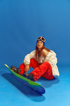 Happy woman in a mix of winter sports attire and summer wear, enjoying snowboarding vacation. against blue studio backgroundの写真素材