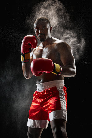 Determined African man, muscular athlete in boxing gear, focused and prepared for fight isolated on black background with dustの写真素材