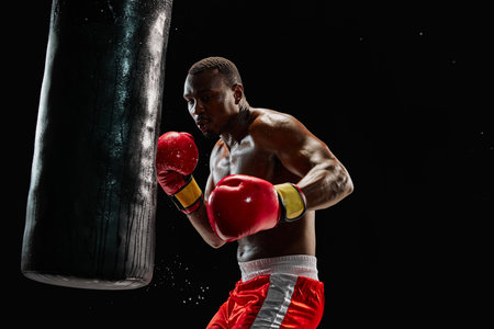 Muscular African man, focused boxer in red gloves delivering powerful punch to a heavy bag isolated on black backgroundの写真素材