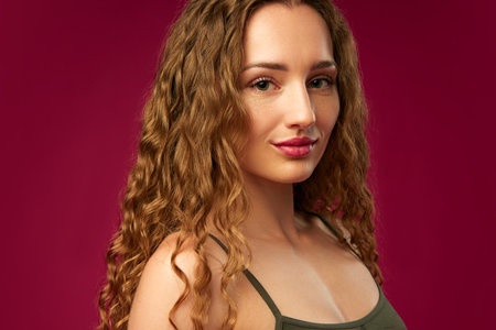 Portrait of young girl with curly hair, nude makeup look posing against magenta studio background....