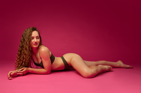 Beautiful young girl with curly long hair posing in underwear, posing on floor against magenta studio backgroundの写真素材