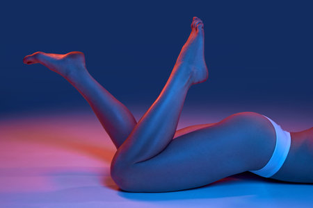 Cropped image of slender, smooth female legs. Model posing in underwear against blue studio background in neon lightの写真素材