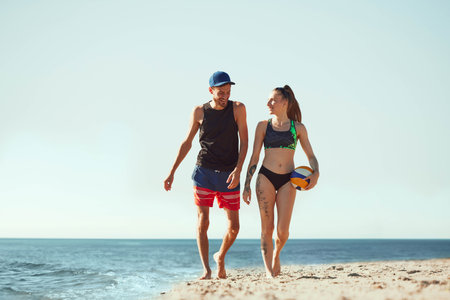 Two young people, man and woman in sportswear walking on beach by the sea with volleyball ball, ready to play on sunny early morning.の写真素材