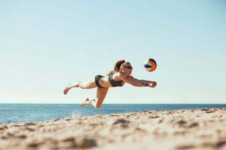 Young girl in sportswear and sunglasses, athlete in motion, playing beach volleyball under sunny clear sky, by the sea. Hitting ball and falling downの写真素材