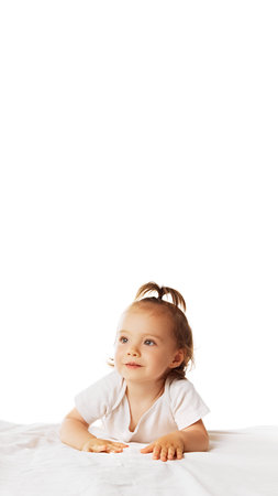Vertical image of adorable little girl, baby in bodysuit, with ponytail leaning on bed and looking isolated on white backgroundの写真素材