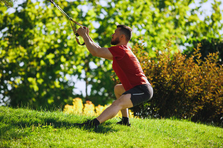 Athletic man with song muscular body, in sports clothing training legs with trx fitness straps outdoors against greenery parkの写真素材