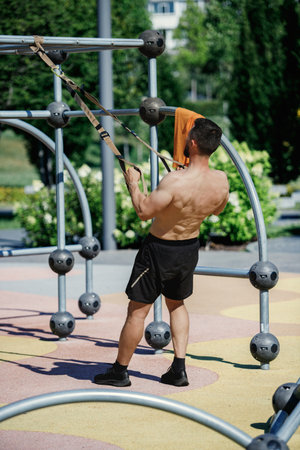 Shirtless man using suspension straps for exercise in outdoor fitness park, wearing black shorts....