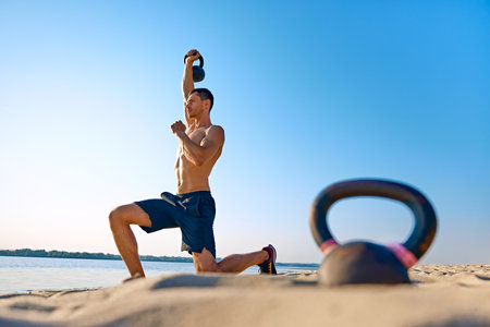 Shirtless man in black shorts performing kettlebell exercise on sandy beach, under clear sunny sky in the background.の写真素材