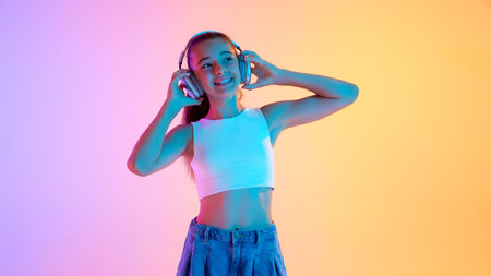 Portrait of beautiful teen girl in white top listening to music in headphones and smiling against gradient pink yellow background in neon lightの写真素材