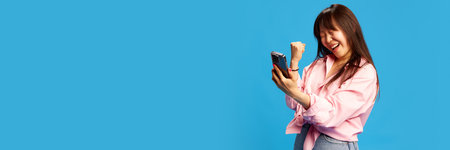 Success, betting, online shopping. Happy Korean girl expressing success while looking on mobile phone screen against blue studio backgroundの写真素材