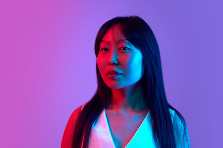 Portrait of confident Asian woman in white shirt posing with serious and calm expression against purple background in neon lightの写真素材