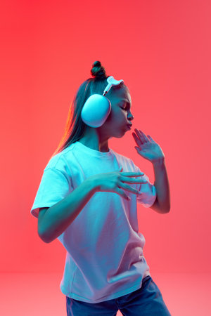 Portrait of young girl, child in casual clothes, t-shirt and jeans, listening to music in headphones and dancing against pink background in neon lightの写真素材