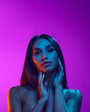Sensual beauty, aesthetics. Attractive young woman with black hair, well-kept skin, plump lips against purple background in neon lightの写真素材