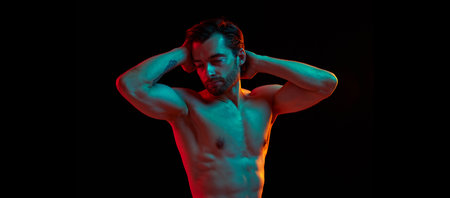 Handsome man with well-kept skin and hair, showing perfect muscular body, posing shirtless against black studio background in neon lightの写真素材