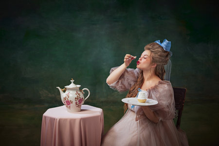 Portrait of beautiful young woman in elegant period attire sitting at table and eating cherry from cake against vintage green backgroundの写真素材