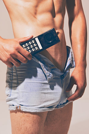 Cropped i,age of relief male torso, muscular tanned shirtless body, model in blue jeans shorts standing with old-style mobile phoneの写真素材