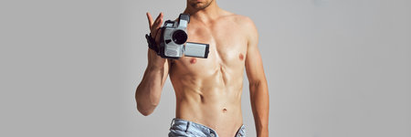 Retro Tech. Cropped image of shirtless man with muscular, athletic body, in denim short standing with vintage camcorder and filmingの写真素材