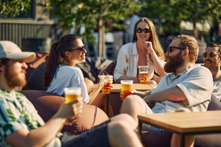 Lively Picnic. Young people, men and women, friends gathering on outdoor restaurant space, cheerfully talking and drinking beer.の写真素材