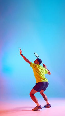 Full-length image of young man, badminton player, athlete serving shuttlecock with racket, training against gradient blue pink background in neon lightの写真素材