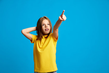 Young girl, teen in yellow polo shirt standing with emotive face and pointing upwards with finger against blue studio background. Look up.の写真素材