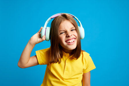 Portrait of young smiling 9 years old girl in yellow polo shirt using headphones to listen to music against blue studio backgroundの写真素材