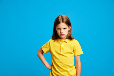 Portrait of teen girl in yellow polo shirt standing with serious face, expressing dissatisfaction against blue studio backgroundの写真素材