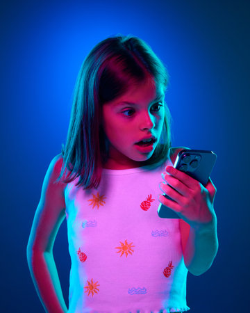 Emotional young girl looking on mobile phone screen with shocked face against blue studio background in neon light. Shock, surprise, sales, newsの写真素材
