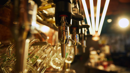 Close-up of multiple shiny beer taps with handles and mirrors, set against blurred background with rows of empty glasses and bar counter in the background.の写真素材