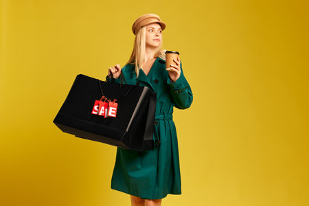Elegant young woman in green trench coat carrying black shopping bags and drinking coffee against yellow studio backgroundの写真素材