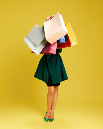 Full-length image of woman in elegant dress and heels holding many colorful shopping bags against yellow studio backgroundの写真素材