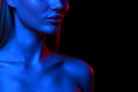 Cropped image of beautiful young woman, shoulders, demonstrating perfect facial lines against black studio background in neon lightの写真素材