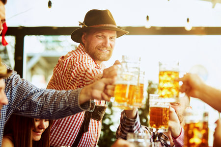 Cheerful bearded man in checkered shirt and hat, cheerful clinking glass of lager beer in celebratory toast, having fun with friends at pubの写真素材