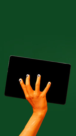 Hand with quirky Halloween themed nails, holding tablet with blank screen against yellow background. News, sales, announcementの写真素材