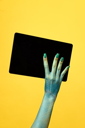 Hand with blue, scaly skin and green nails holds black tablet device with blank screen facing viewer against yellow background. Social media blog post, newsの写真素材