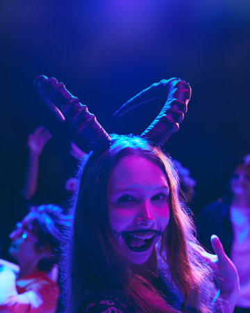 Portrait of smiling young girl with creepy makeup and animal horns on head celebrating Halloween...