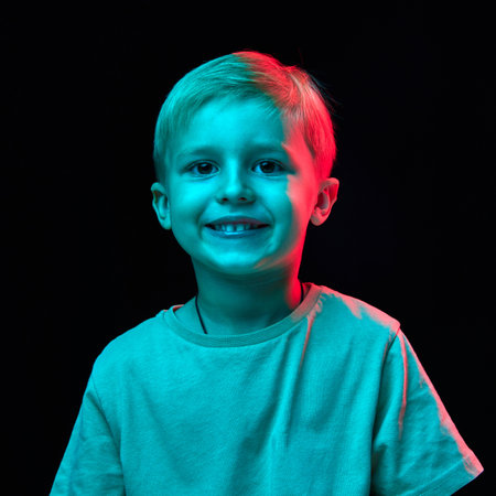 Portrait of little 4 years old boy with blonde hair, standing with cheerful smile, wearing t-shirt and posing against black studio background in neon lightの写真素材